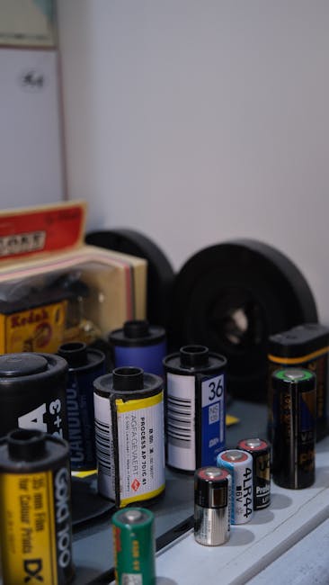 A close-up view of a collection of various batteries, including AA, AAA, C, D, and 9V types, arranged on a flat surface indoors. Some batteries are standing upright, while others are lying on their sides. In the background, partially visible, are black speakers, a cardboard box, and a white appliance or cabinet. The lighting appears natural, coming from an indoor setting, with no visible packaging materials or furniture directly related to house removals. The scene reflects typical household items that might be packed or prepared for a home relocation, aligning with the services offered by Man and a Van Ilford. This detailed visual emphasizes proper handling, packing, or transporting of small household items during a furniture transport or packing and moving process.