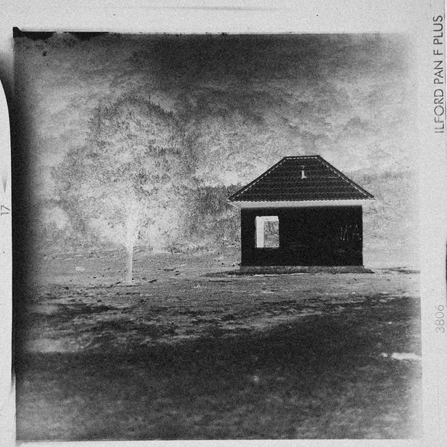 Black and white photograph of a small, single-story house with a pitched roof, positioned in a remote, open area. The house has a large rectangular window on the front wall and no visible door, possibly under construction or renovation. Surrounding the house is an expansive landscape with sparse, leafless trees and uneven ground, suggesting a rural or undeveloped setting. The sky appears overcast, casting diffuse lighting over the scene. In the foreground, there are scattered rocks or debris, indicating a rough, unpaved surface. The image was taken outdoors, with the house centered in the composition, highlighting a quiet, isolated environment. This scene can relate to home relocation or transport services, as part of the process of moving or packing furniture and belongings to or from a distant property. Occasionally, [COMPANY_NAME] may handle such moving tasks, including logistics of loading and transporting items in rural or less accessible areas.