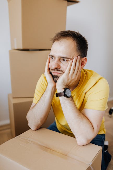 A man with short brown hair and a beard, wearing a yellow t-shirt and a black wristwatch, is sitting at a table in a room filled with cardboard moving boxes. He rests his head in his hands with a worried expression, suggesting stress during a home relocation process. Behind him, several stacked cardboard boxes and a doorway are visible, indicating an interior space prepared for furniture transport and packing. The scene captures the common challenges faced during last-minute house removals, illustrating the need for professional moving services like those offered by Man and a Van Ilford, who specialise in furniture transport, packing, and loading processes essential to resolving last-minute issues in house relocations.