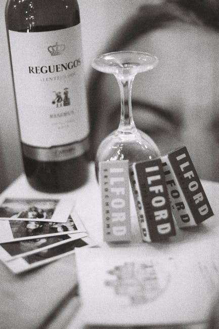 A partially filled wine bottle labeled 'Reguengo' positioned on the left side of the image, next to a clear, upside-down wine glass on its stem, with the bowl facing downward, centrally placed on a flat surface. Several small printed photographs or postcards are spread out in front of the bottle and glass, with some overlapping. In the background, there is a finger or hand holding three small labels or tags, each emblazoned with 'ILFORD' and 'LIFE FORD' in bold capital letters. The surface appears to be indoors and well-lit, and the arrangement suggests a casual presentation of items associated with a house or apartment, possibly related to packing or organizing during a move. The image captures a moment where drinkware, printed materials, and promotional tags are grouped together, with no further context visible about the environment or activity.
