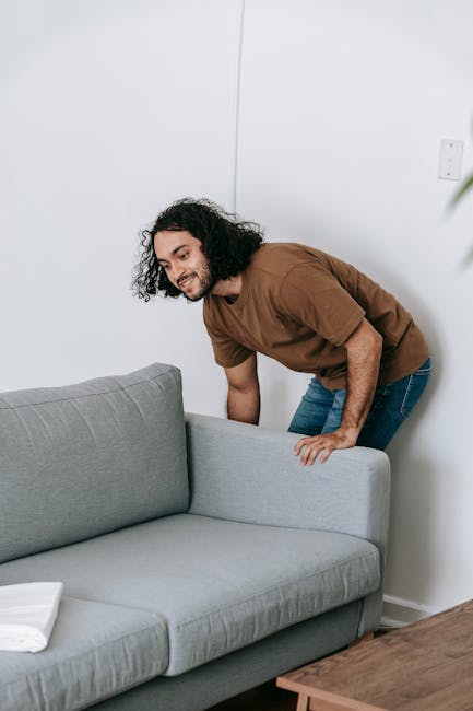 A man with curly dark hair, wearing a brown t-shirt and blue jeans, is in the process of preparing a home for a furniture removal. He is bending over slightly, lifting or adjusting the grey fabric-upholstered sofa, which is positioned against a white wall with an electrical socket visible. The room appears to be a living area with a wooden side table nearby. This scene captures the initial stages of packing and moving, illustrating the careful handling of furniture by a member of a removals team, possibly from Man and a Van Ilford, as they assist with home relocation or furniture transport inside the property in preparation for loading onto a vehicle for transportation.