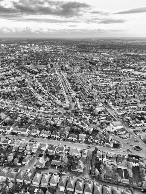 A black-and-white aerial photograph shows a densely populated residential neighbourhood with rows of terraced houses, semi-detached homes, and small apartment buildings. Streets and avenues run through the area, some with visible parked cars along curbs. In the foreground, a wide street features a roundabout. To the right of this street, a large open space or backyard is visible, surrounded by additional housing. The middle ground displays a mix of smaller streets and larger roads, some lined with trees, with utility poles and streetlights visible along the roadways. In the background, the landscape extends to the horizon with more urban development, and the sky above is partly cloudy with patches of sunlight breaking through. The image contextually supports house removals and moving logistics by depicting a typical residential area where furniture transport and home relocation services by Man and a Van Ilford are applicable, especially during packing, loading, and transportation phases of a move in the IG1 postcode area.