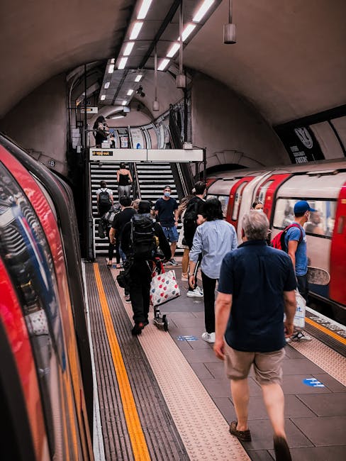 Inside Ilford Station, a group of passengers is disembarking from a red and grey train on an underground platform. The platform features textured yellow and grey safety lines, with passengers carrying luggage, backpacks, and shopping bags. Some individuals are walking toward the station stairs, where a man with a backpack is ascending the steps. The station ceiling is curved, with modern lighting fixtures and suspended cables visible. On the right side, the train doors are open, revealing the interior, while on the left, part of another train is visible. The scene captures a typical busy station environment, which Man and a Van Ilford might consider during home relocation or furniture transport logistics involving moving through public transport hubs.