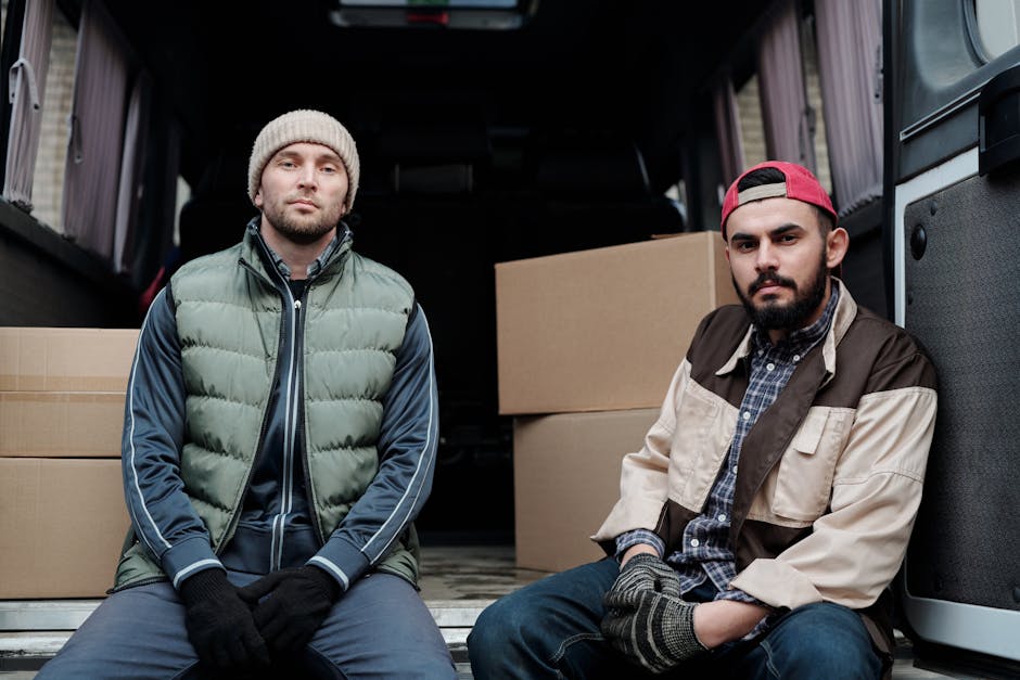 Two men are engaged in a home relocation task outside a residential property, loading a large cardboard box into a white commercial van parked on a brick driveway. One man, wearing a black quilted ves
