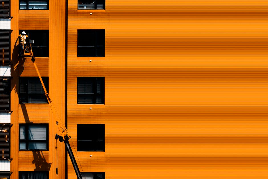 A worker wearing a white safety helmet and high-visibility vest is positioned on a hydraulic lift attached to a black cherry picker truck, accessing the exterior of a multi-storey residential building with orange corrugated metal cladding. The worker appears to be engaged in a home relocation task, possibly loading or unloading items through one of the open windows. The building's windows are dark, and the hydraulic lift's extendable arm is angled upward, supporting the worker's platform. Inside the distance of the scene, the ground is not visible, indicating the lift is extended at a significant height. This image demonstrates the use of professional equipment, such as the hydraulic lift, for furniture transport and packing during house removals, aligning with services offered by Man and a Van Ilford, especially when lift delays impact load handling in an urban environment.