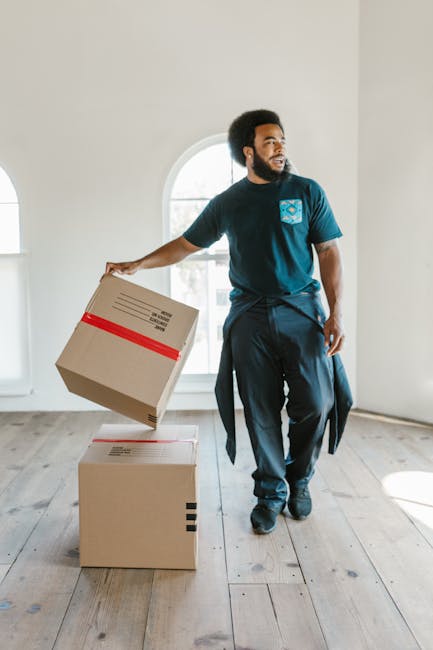 A man with dark skin, curly black hair, and a beard is inside a bright, spacious room with white walls and large arched windows allowing natural daylight. He is wearing a navy t-shirt with a logo on the chest and dark trousers, and is in the process of moving or packing. He is holding a cardboard box with red tape, preparing to place it on top of another similar box on the light wooden floor. The room has minimal furnishings and appears to be in the midst of a home relocation. Man and a Van Ilford’s professional furniture transport and packing services are implied by the scene, reflecting careful handling and organized packing during a house move, which may encounter delays affecting timing and logistics, as suggested by the page title about lift delays during removals jobs.
